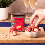 A tin of Cocoba Raspberry Champagne Truffles sits on a wooden board next to a small bowl of the truffles, with a hand reaching for one. Two champagne flutes and a potted plant are blurred in the background.