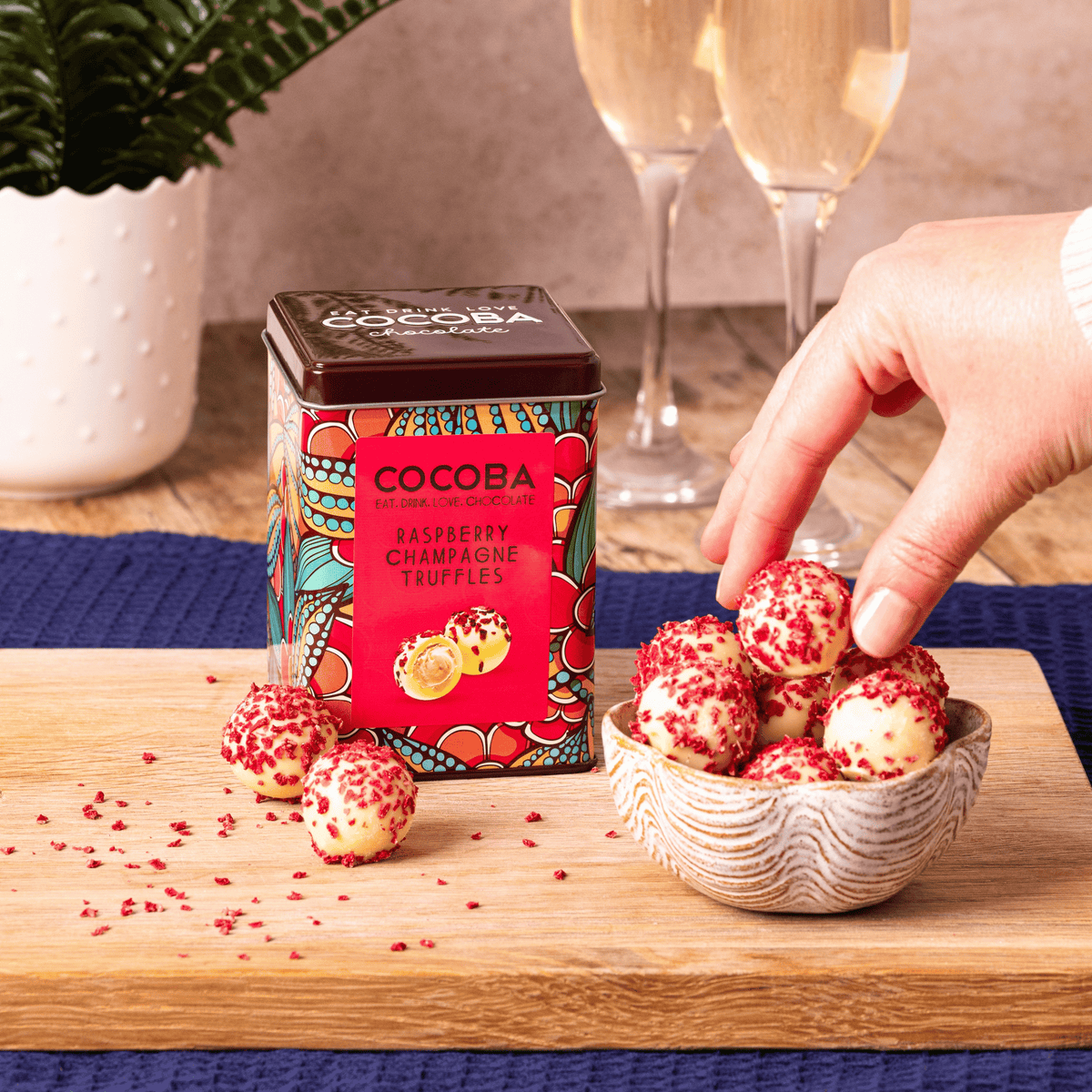 A tin of Cocoba Raspberry Champagne Truffles sits on a wooden board next to a small bowl of the truffles, with a hand reaching for one. Two champagne flutes and a potted plant are blurred in the background.