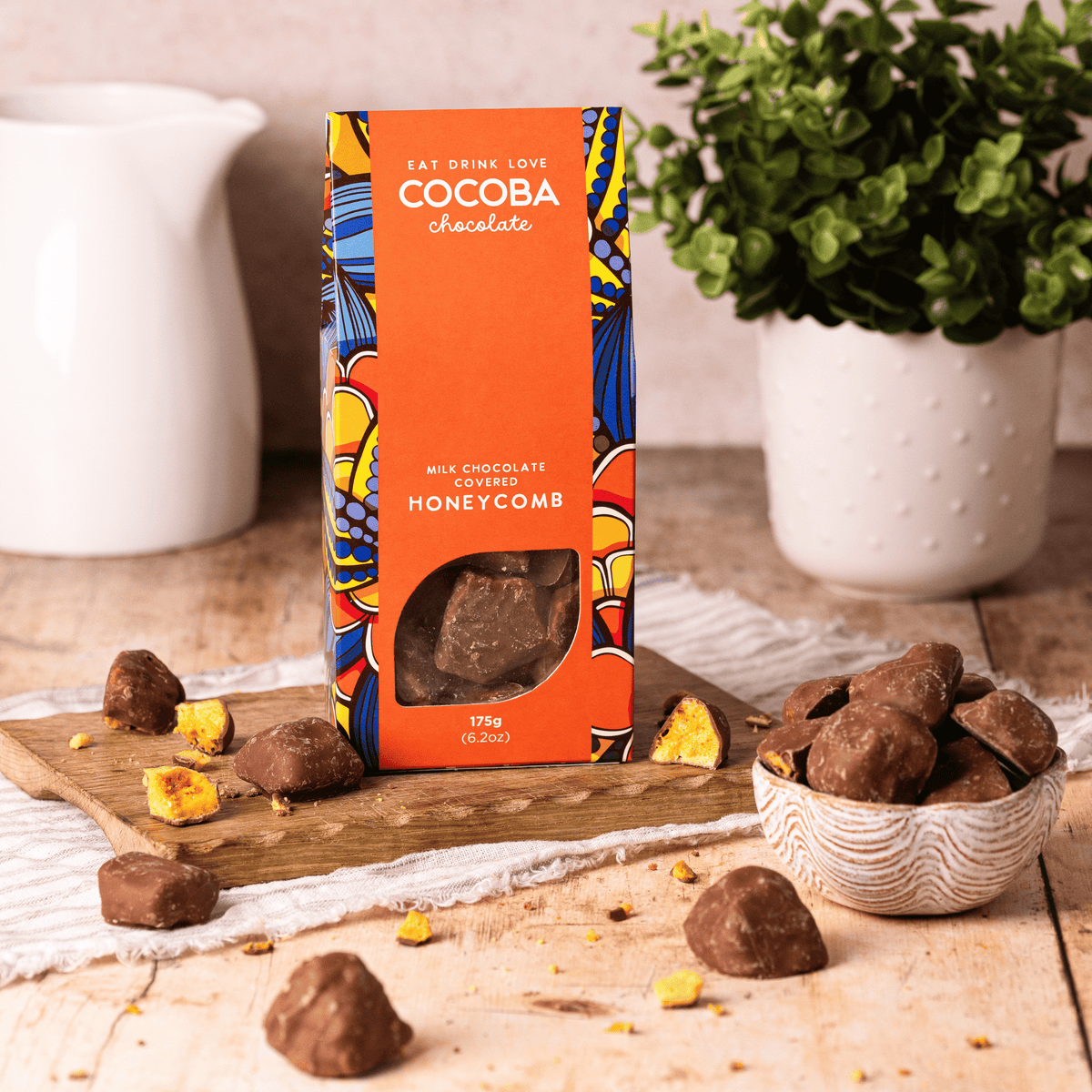 Cocoba chocolate honeycomb packaging on a wooden board with chocolates and a plant in the background.