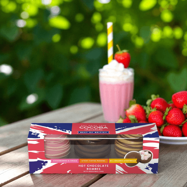 Box of hot chocolate bombs with a strawberry milkshake and strawberries on a wooden table outdoors.