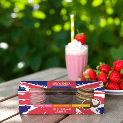 Box of hot chocolate bombs with a strawberry milkshake and strawberries on a wooden table outdoors.