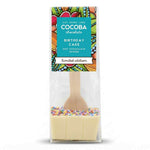A packaged chocolate hot chocolate spoon shaped like a birthday cake, with a wooden spoon handle, white chocolate base, colorful sprinkles, and a decorative label reading COCOBA chocolate.