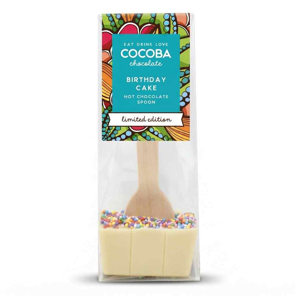 A packaged chocolate hot chocolate spoon shaped like a birthday cake, with a wooden spoon handle, white chocolate base, colorful sprinkles, and a decorative label reading COCOBA chocolate.