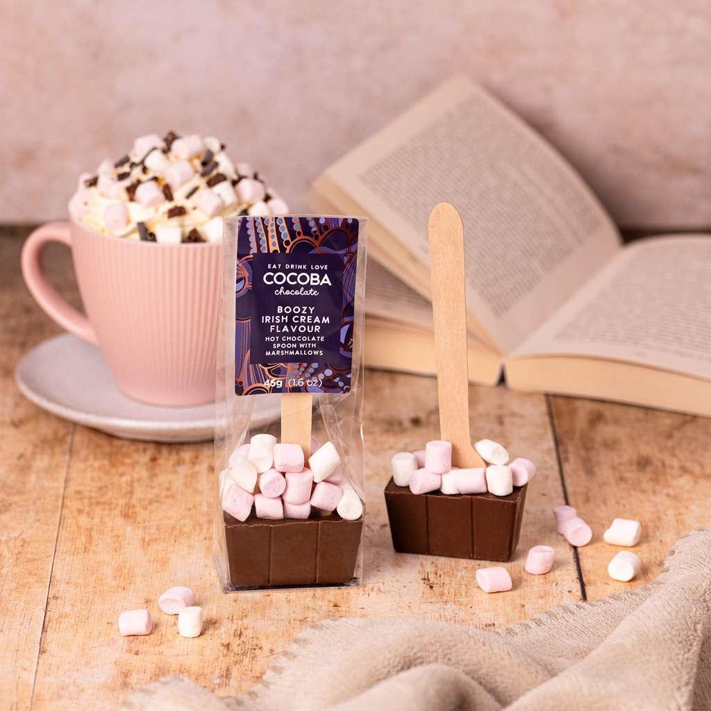 Cocoba Irish cream flavoured milk chocolate hot chocolate spoon with marshmallows, next to a mug of hot chocolate and an open book on a wooden surface.