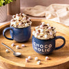 Two blue mugs from Cocoba labeled 'CHOCOHOLIC', with hot chocolate and marshmallows on a wooden tray.