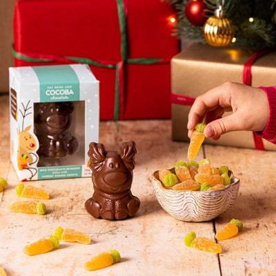 Chocolate reindeer figure, with jelly carrot sweets, and a child's hand reaching for a jelly carrot sweet with Christmas decorations around.