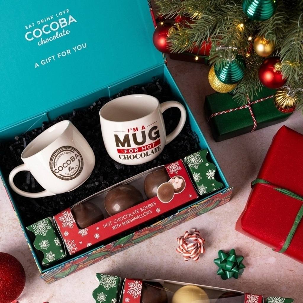 A Christmas-themed gift set in an open teal box featuring two white mugs, one with a COCOBA chocolate logo and the other reading 'I'M A MUG FOR HOT CHOCOLATE,' along with a box of hot chocolate bombs and marshmallows. The scene is styled with a decorated Christmas tree, wrapped presents, and candy on a light pink surface.