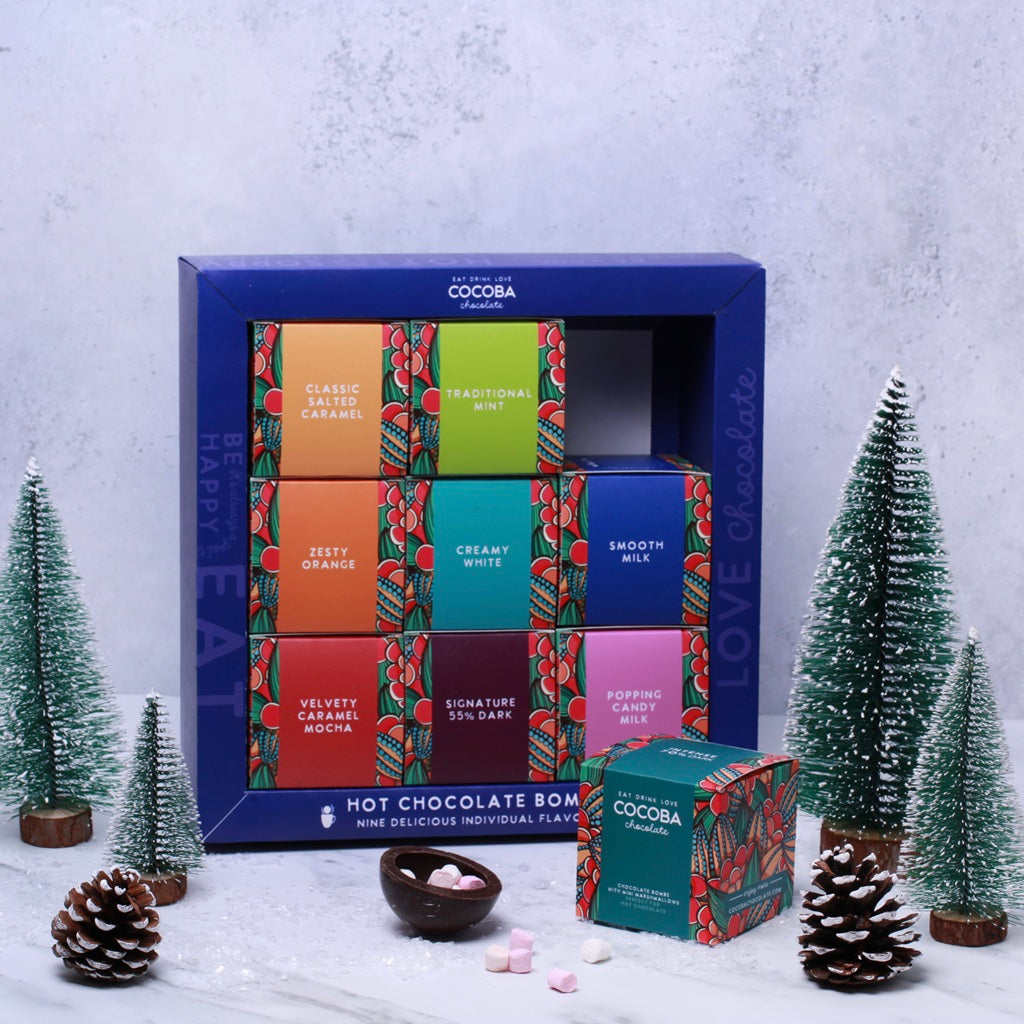 Cocoba Christmas Hot Chocolate Bombe Selection Box with colorful packaging on a snowy background with decorative trees and pinecones. 