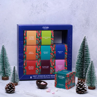 Cocoba Christmas Hot Chocolate Bombe Selection Box with colorful packaging on a snowy background with decorative trees and pinecones. 