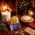 A bag of cocoa chocolate mini carrot jelly sweets is displayed on a rustic wooden table with a warm holiday scene. The background features a burning fireplace, a glass of milk, a star-topped pie, pinecones, and a handwritten note that reads 'For Santa and his Reindeer'.