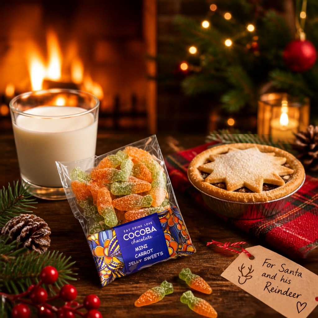 A bag of cocoa chocolate mini carrot jelly sweets is displayed on a rustic wooden table with a warm holiday scene. The background features a burning fireplace, a glass of milk, a star-topped pie, pinecones, and a handwritten note that reads 'For Santa and his Reindeer'.