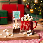 Hot chocolate spoon with star-shaped mini-marshmallows Christmas packaging next to a mug with Christmas design, hot chocolate, and star marshmallows on a wooden board with a festive background.