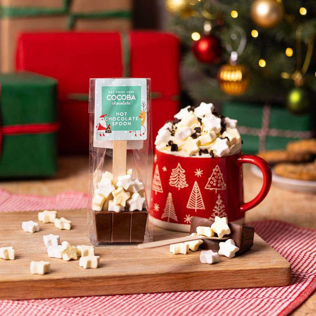 Hot chocolate spoon with star-shaped mini-marshmallows Christmas packaging next to a mug with Christmas design, hot chocolate, and star marshmallows on a wooden board with a festive background.
