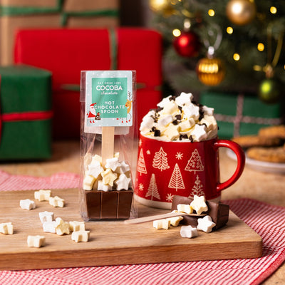 Hot chocolate spoon with star-shaped mini-marshmallows Christmas packaging next to a mug with Christmas design, hot chocolate, and star marshmallows on a wooden board with a festive background.