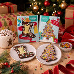 Two Christmas-themed Chocolate Bars in Festive boxes displayed on a wooden table. The left box contains a stocking-shaped Chocolate Bar with white icing and red/green sprinkles, and the right box contains a tree-shaped Chocolate Bar covered in multicolored candy.