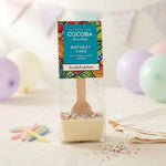 A packaged hot chocolate spoon with a colorful 'Cocoba Chocolate' label, filled with white chocolate and topped with rainbow sprinkles, set on a table with pastel balloons and candles in the background.