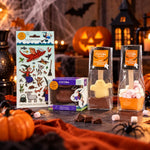A Halloween-themed chocolate gift bundle displayed on a rustic table with pumpkins, candles, and spider decorations. The set includes a sheet of Room on the Broom stickers, and Halloween themed hot chocolate bombes and spoons.
