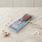 A heart-shaped chocolate pop in its blue and pink packaging, displayed on a light marble surface with scattered mini marshmallows and chocolate shavings.