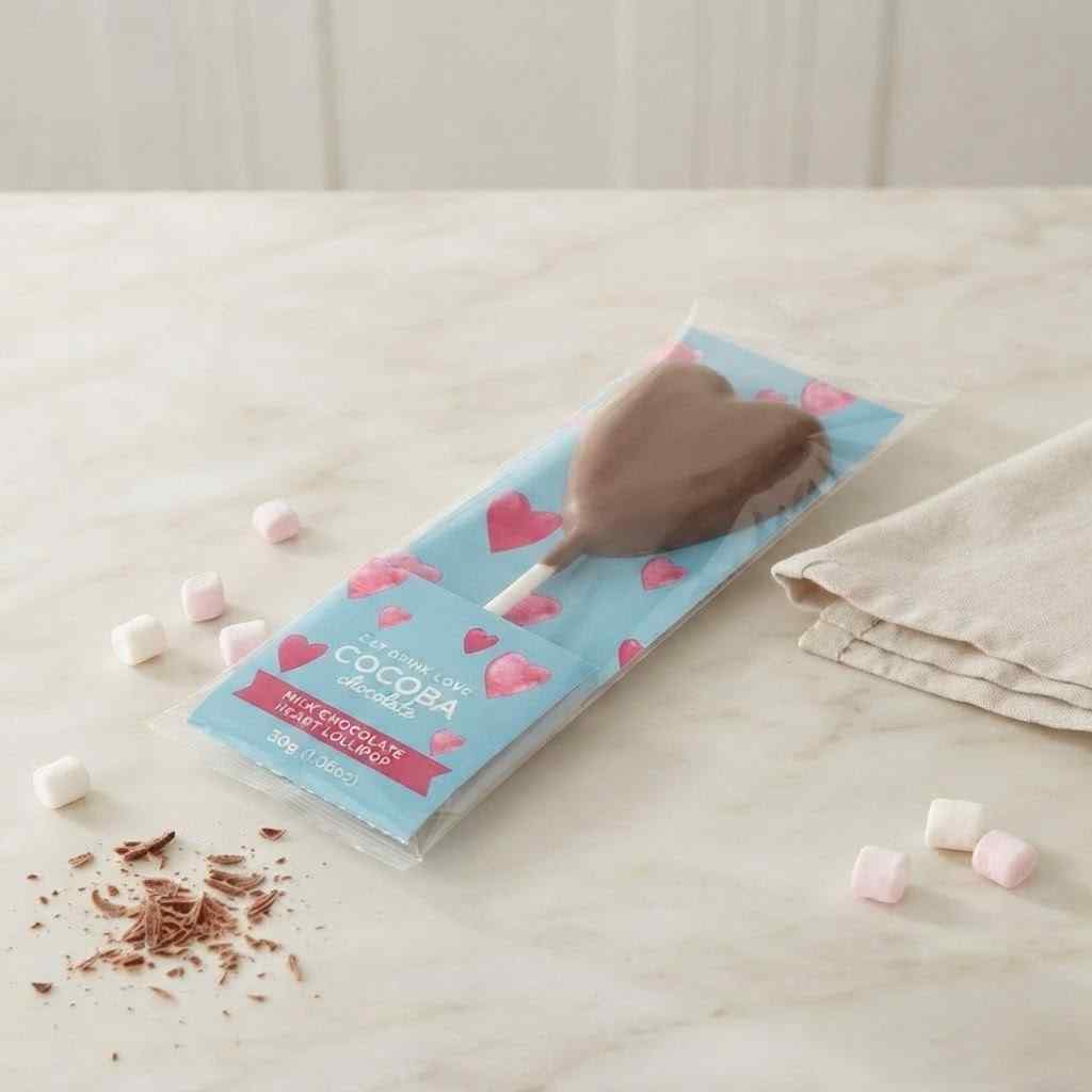 A heart-shaped chocolate pop in its blue and pink packaging, displayed on a light marble surface with scattered mini marshmallows and chocolate shavings.