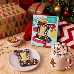 A boxed Christmas stocking chocolate bar in festive Christmas packaging sits on a wooden table beside a Christmas stocking chocolate bar on a plate, with a mug of hot chocolate with marshmallows, wrapped gifts, and a Christmas tree in the background. 