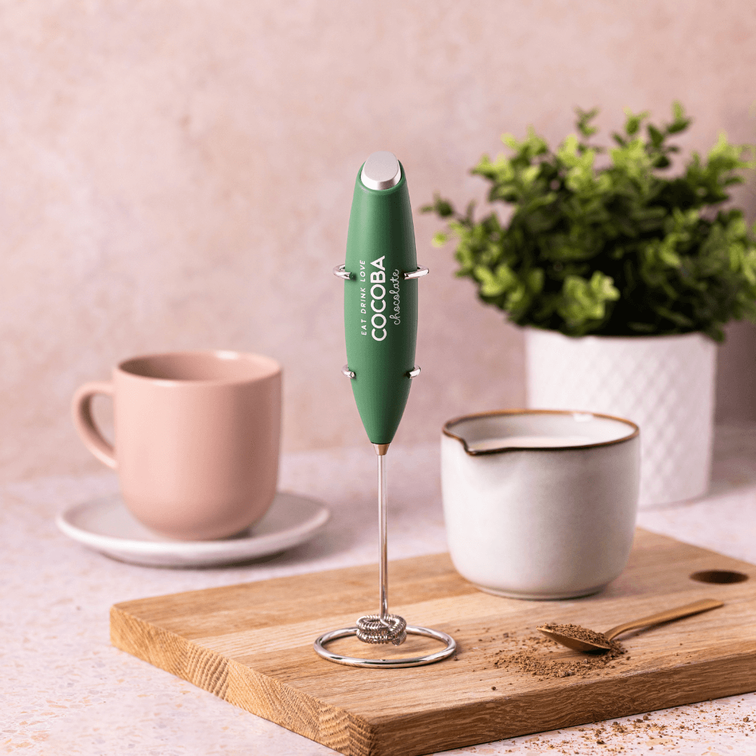 Green Cocoba milk frother for making a smooth and creamy Hot Chocolate, on a wooden board with a cup, saucer, and plant in the background.