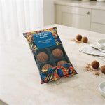 A package of chocolate-covered caramel truffles on a light marble countertop with a few loose truffles and cocoa powder scattered nearby.