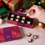 A hand holds a star-shaped chocolate truffle from an open box of Cocoba chocolates, with the matching patterned lid beside it. The scene is styled with for Christmas with festive wrapped gift boxes, ornaments, and evergreen branches on a light surface.
