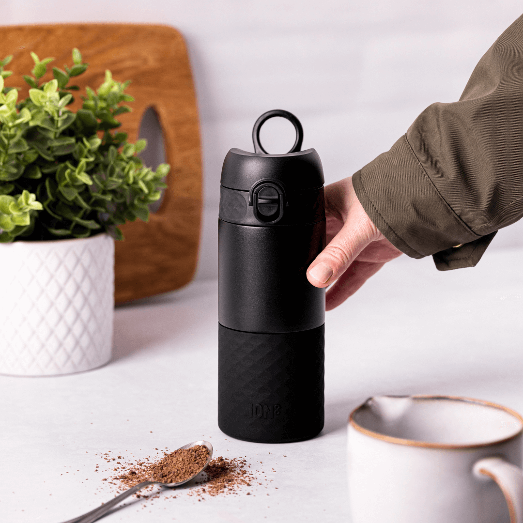 Hand holding a black travel cup with a spoonful of hot chocolate flakes and a jug in the background.