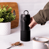 Hand holding a black travel cup with a spoonful of hot chocolate flakes and a jug in the background.