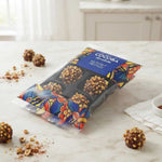 A package of chocolate-covered nuts and dried truffles on a light marble countertop, with a few loose truffles and nut crumbs scattered around. The packaging is colorful with a blue label reading 'COCOBA chocolate' and 'HAZELNUT TRUFFLES'.