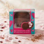 A boxed hot chocolate bomb with marshmallow hearts, shown in its retail packaging on a soft pink surface with scattered marshmallows and cocoa powder.