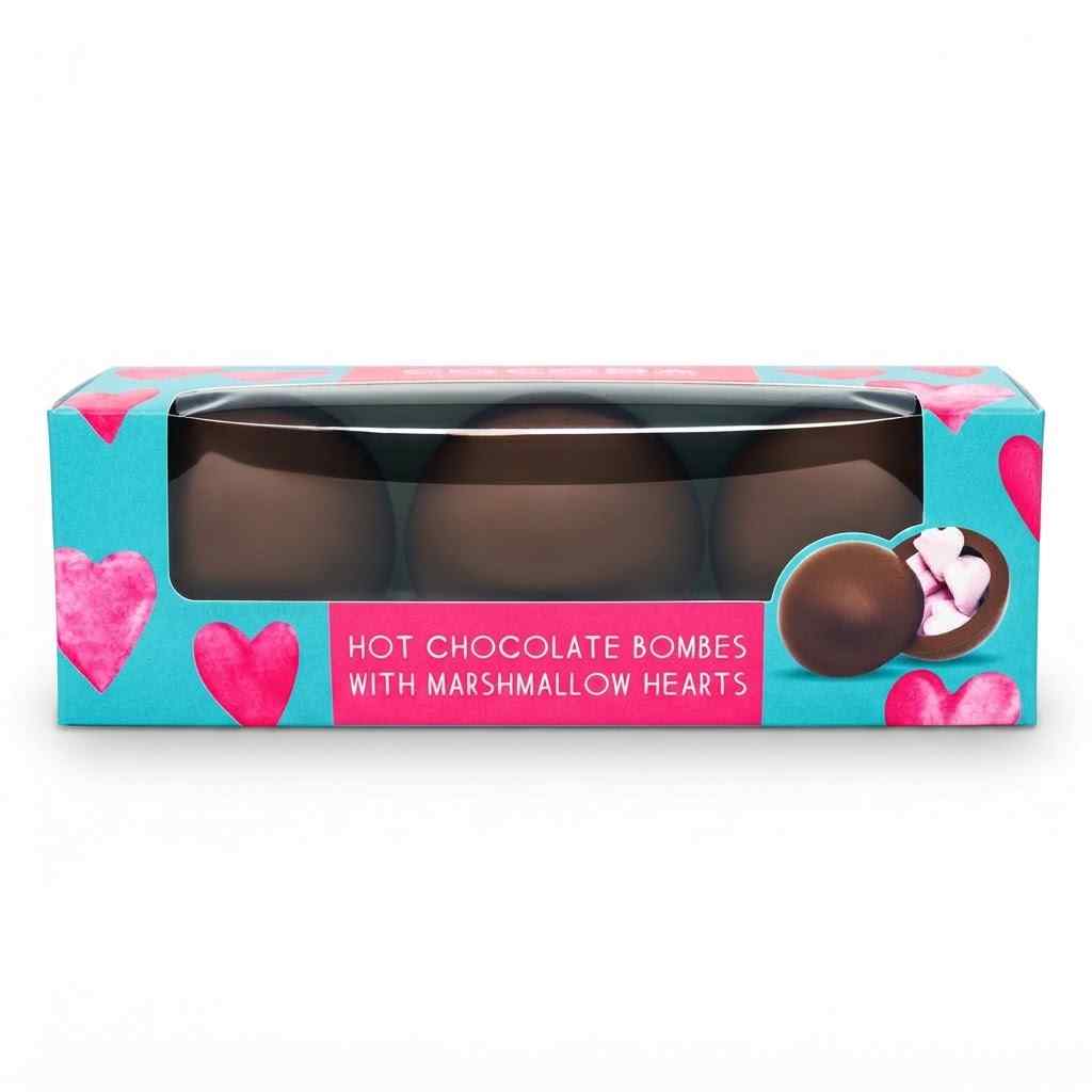 A boxed set of hot chocolate bombs with marshmallow hearts, shown in a turquoise and pink retail package with a clear window revealing three chocolate bombs inside.