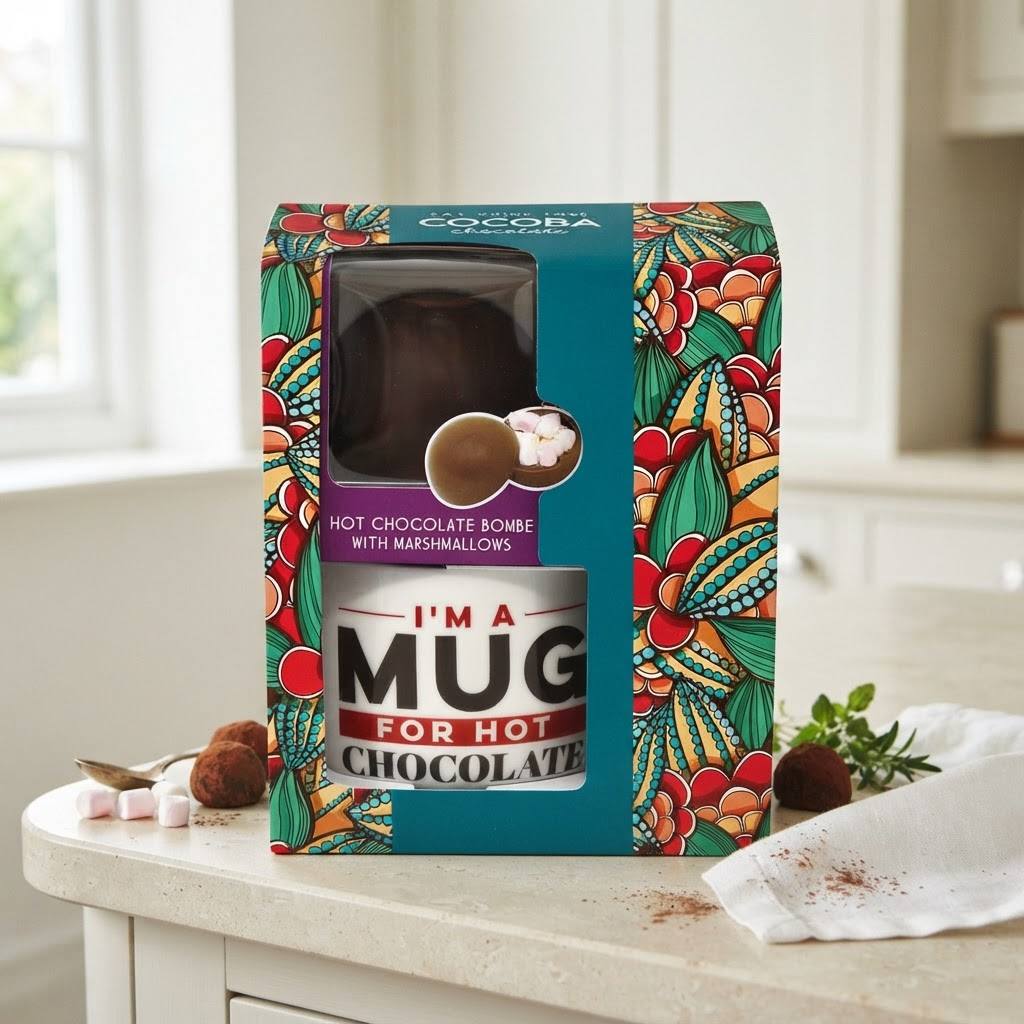 A boxed hot chocolate bomb gift set displayed on a kitchen counter. The packaging is teal with a colorful floral cocoa plant pattern and reads 'I'M A MUG FOR HOT CHOCOLATE' and 'HOT CHOCOLATE BOMB WITH MARSHMALLOWS'.