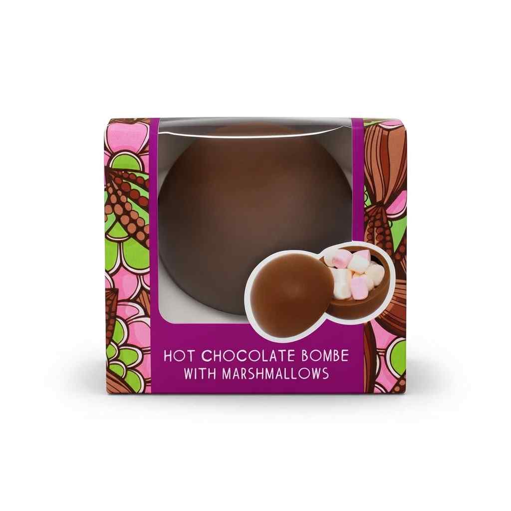 A boxed hot chocolate bombe with marshmallows, shown in its retail packaging on a white background. The package features a clear window displaying a large round chocolate bomb, with an inset showing the chocolate dome cracked open to reveal pink and white marshmallows inside.