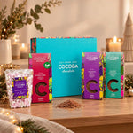 A holiday-themed chocolate gift set from Cocoba Chocolate on a wooden table, featuring a large turquoise box, three colorful chocolate boxes, a bag of marshmallows, and cocoa powder, surrounded by warm candles and festive greenery.