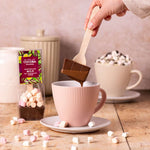 Person adding a hot chocolate spoon into a mug with marshmallows, surrounded by COCOBA Chocolate packaging.
