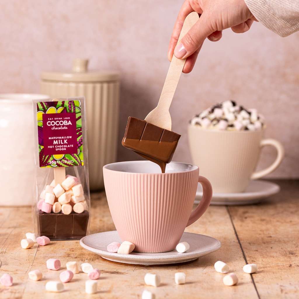 Person adding a hot chocolate spoon into a mug with marshmallows, surrounded by COCOBA Chocolate packaging.
