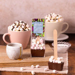 Hot chocolate bomb with marshmallows on a wooden surface with mugs and a package in the background.