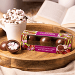 Box of Cocoba hot chocolate bombs with marshmallows on a wooden tray with a cup of hot chocolate in the background.