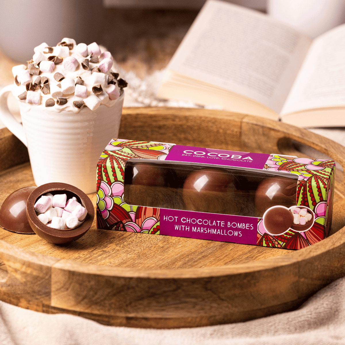 Box of Cocoba hot chocolate bombs with marshmallows on a wooden tray with a cup of hot chocolate in the background.