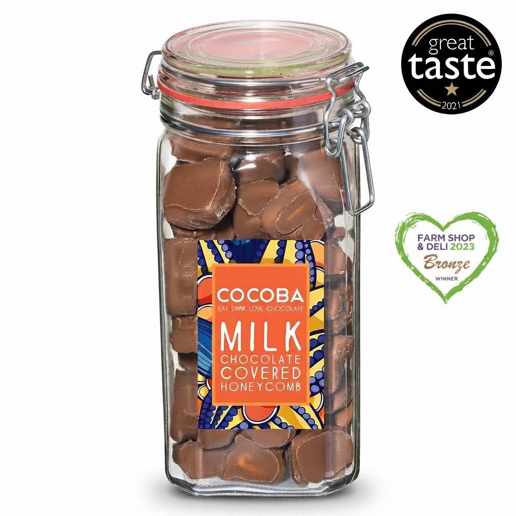 A glass jar of Cocoba Milk Chocolate Covered Honeycomb candies, filled with brown chocolates and featuring a colorful orange, blue, and yellow label. The jar is shown on a white background with a 'great taste 2021' badge on the top right and a 'Farm Shop & Deli 2023 Bronze Winner' heart badge on the bottom right.