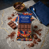 Cocoba chocolate bar packaging on a stone surface with chocolate pieces and a bowl of cacao nibs.