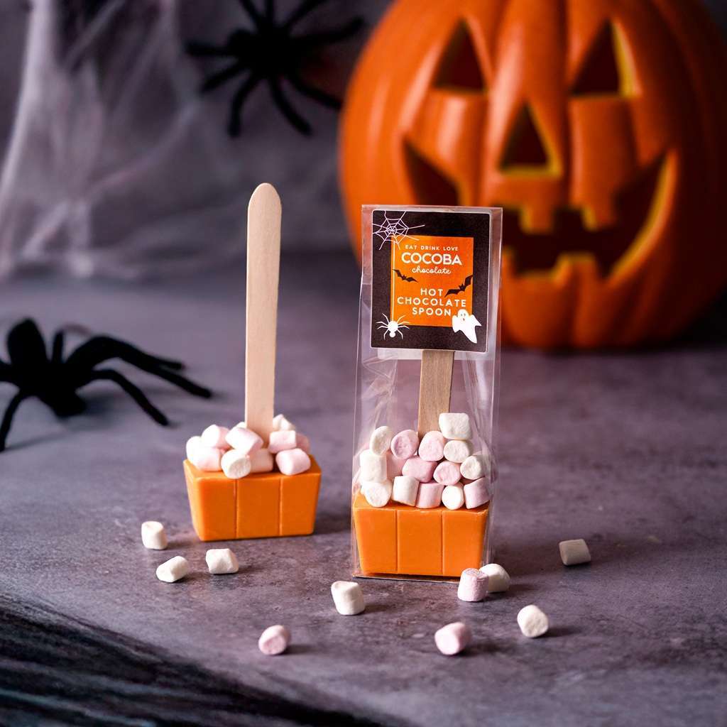 Halloween-themed hot chocolate spoons with marshmallows on a dark surface, featuring a pumpkin and spider decorations.