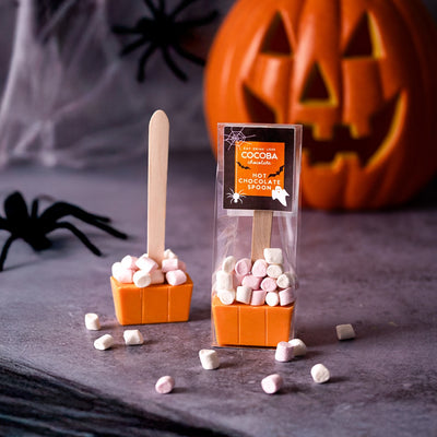 Halloween-themed hot chocolate spoons with marshmallows on a dark surface, featuring a pumpkin and spider decorations.