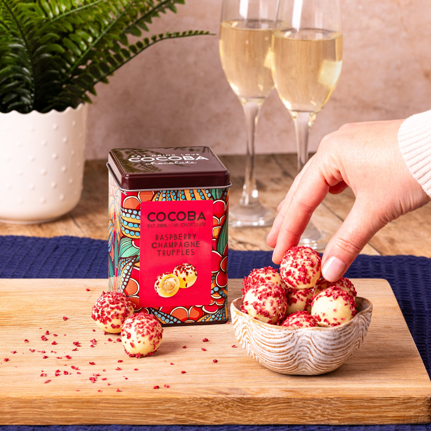 Cocoba raspberry and champagne white chocolate truffles with a person picking one from a bowl on a table, and two glasses of champagne in the background.