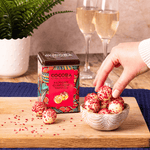 Raspberry and Marc de Champagne Truffles Gift Tin on a wooden board with two glasses of champagne in the background. 