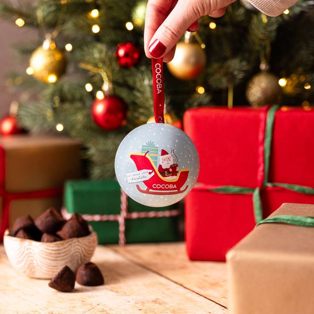 COCOBA Salted Toffee Truffle Christmas Bauble held by a hand with a festive background of presents and a tree.
