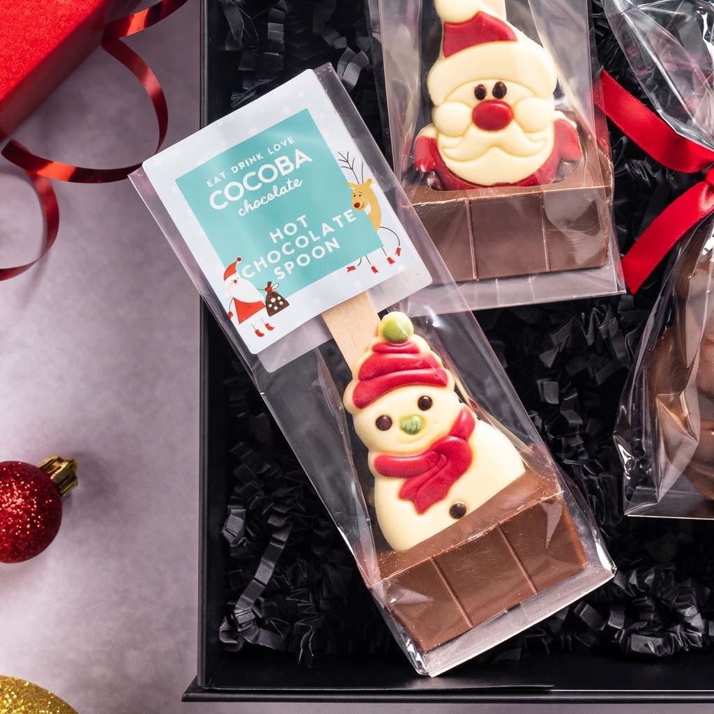 A gift box of chocolate lollipops featuring a snowman and Santa Claus design, with a package labeled 'COCOPA chocolate HOT CHOCOLATE SPOON' on a gray background with holiday decorations.