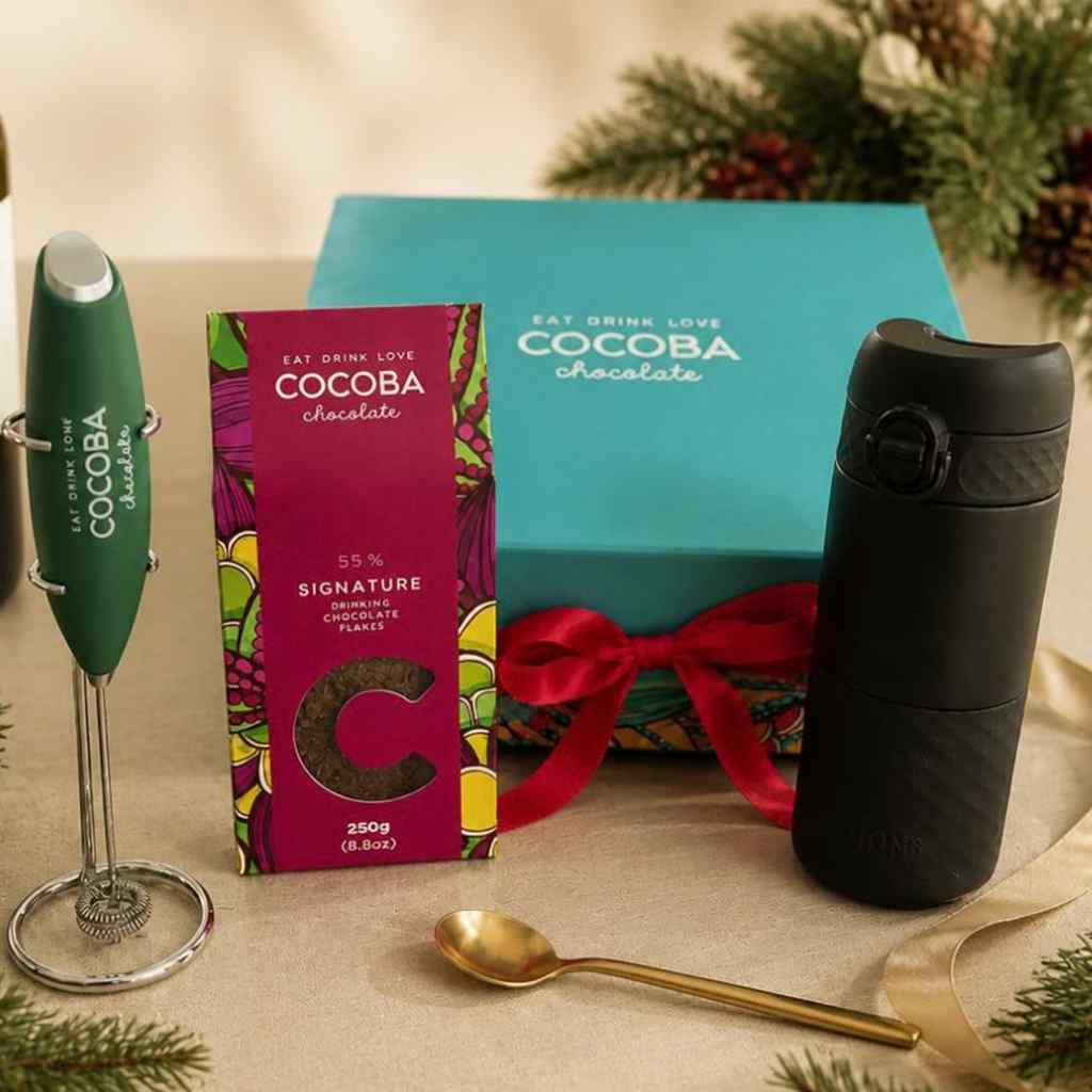 A Cocoba chocolate gift set featuring a green Cocoba Milk frother, a box of signature drinking chocolate flakes, and black travel mug, with a turquoise Cocoba gift box, and festive greenery in the background.