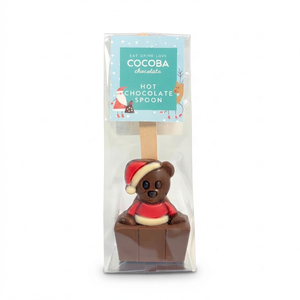 A packaged chocolate hot chocolate spoon featuring a teddy bear wearing a Santa hat and red sweater, sitting in a small chocolate box. The item is in clear retail packaging with a printed label that reads 'COCOBA chocolate HOT CHOCOLATE SPOON'.